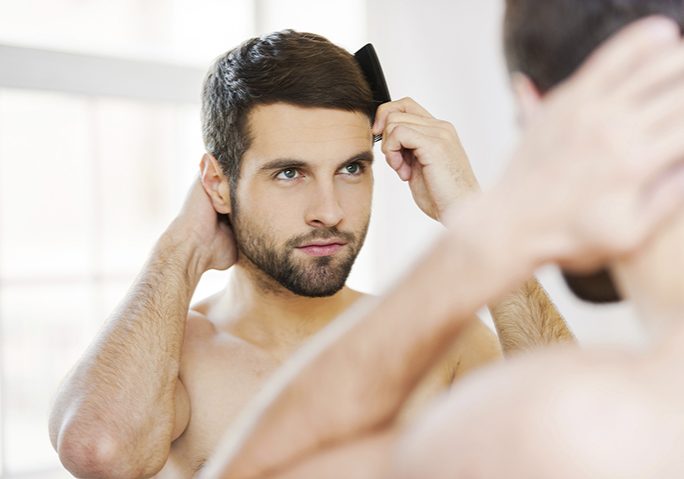 Morning routine. Rear view of handsome young beard man combing his hair while standing against a mirror